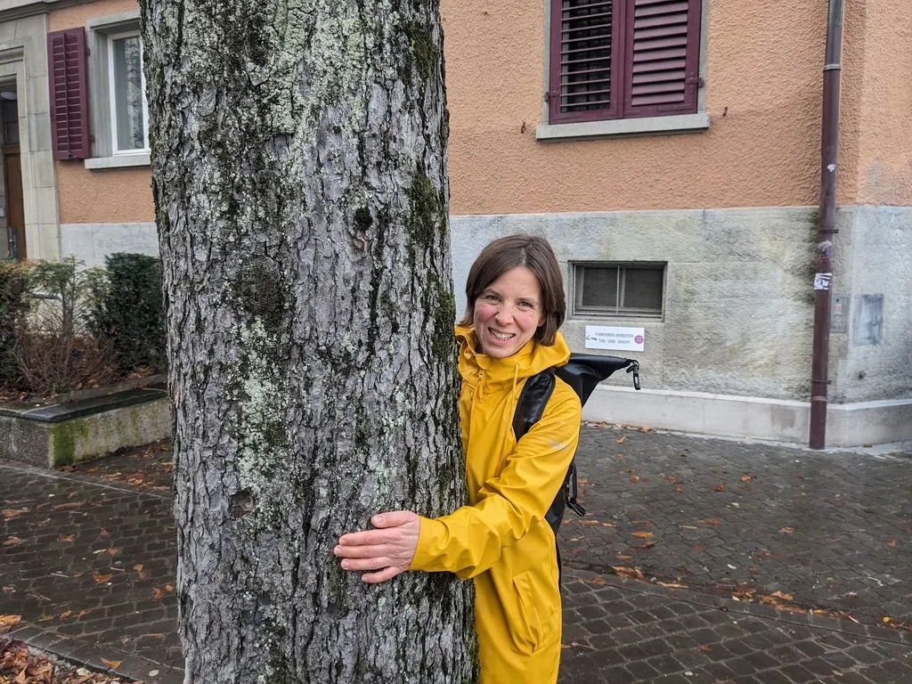 Lachende Frau in gelbem Mantel umarmt Baum.