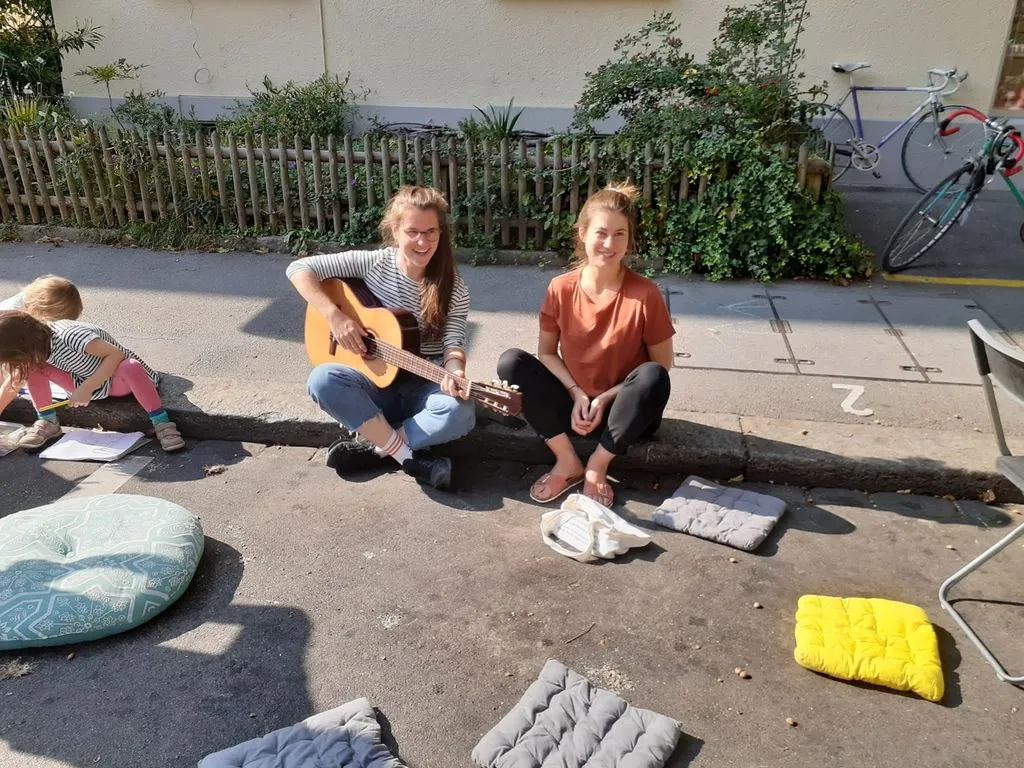 Zwei junge Personen mit Gitarre sitzen auf Kissen auf Gehsteig.